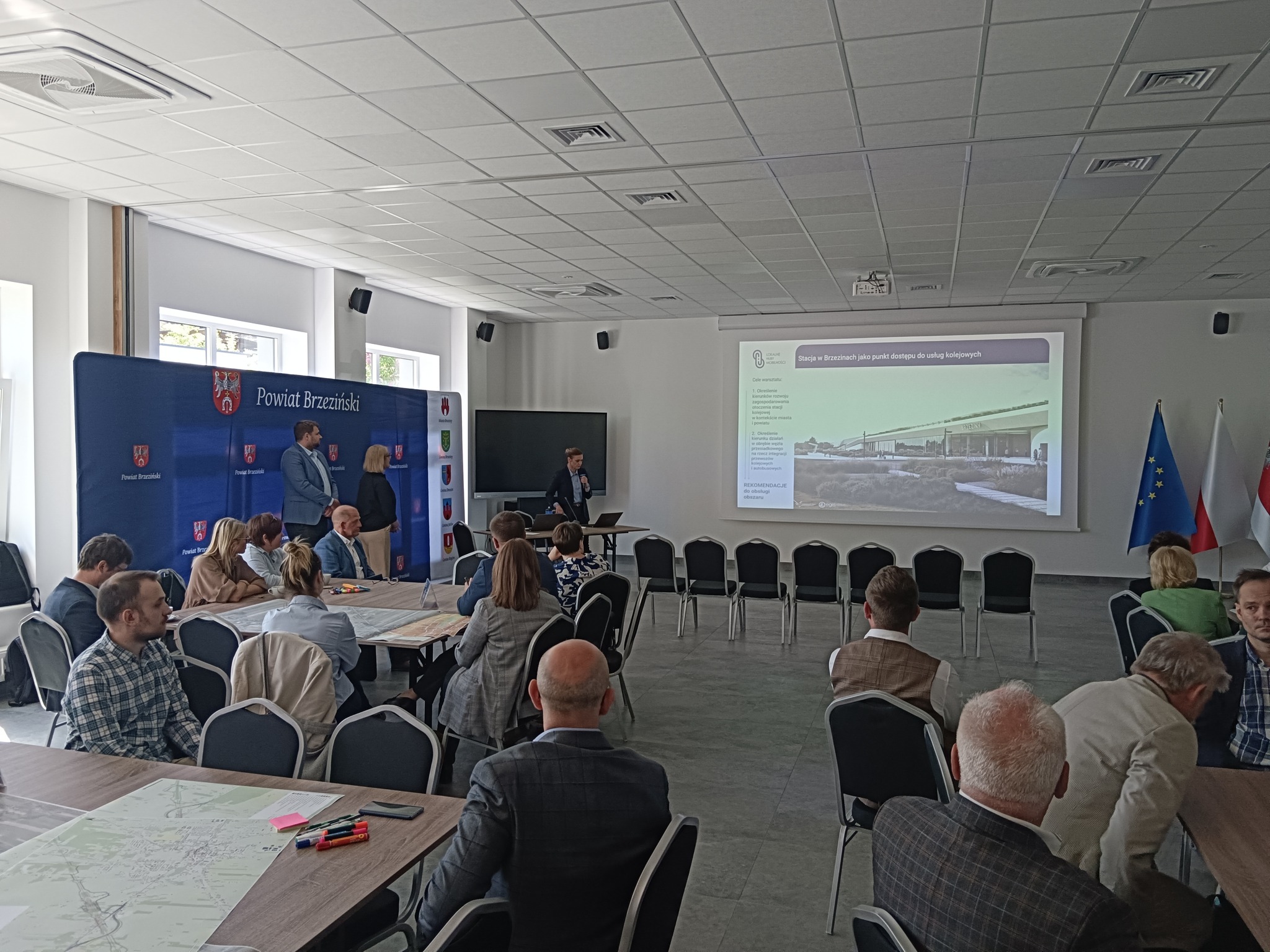 Zdjęcie przedstawiające spotkanie na sali konferencyjnej, w tle zdjęcia widać wyświetlaną na planszy prezentację przedstawiającą wizualizacje dworca PKP, obok po prawej flagi UE, Polski i Powiatu Brzezińskiego. Po lewej stronie stoi osoba omawiająca prezentację, dalej widać dwie osoby stojące a tle niebieskiej ścianki z herbem i napisem Powiat Brzeziński. Na pierwszym planie zdjęcia widać osoby siedzące przy stołach, na których leżą mapy z planami CPK.