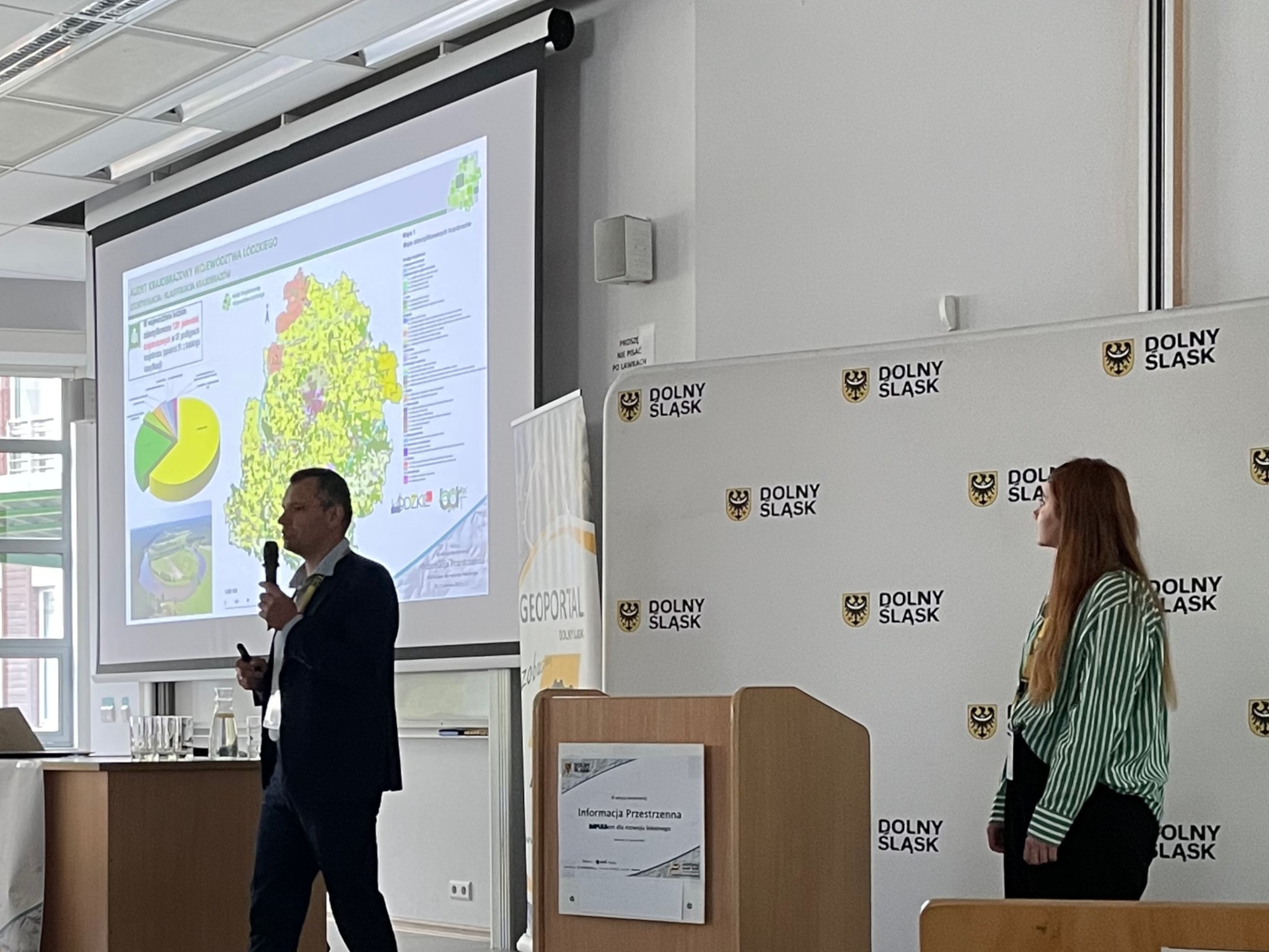 Zdjęcie zXI edycji konferencji "Informacja przestrzenna IMPULSem dla rozwoju lokalnego". Po lewej stronie, obok prezentacji, przodem do widowni stoi Dyrektor BPRWŁ. W tle zdjęcia widać prezentacje z mapą Województwa Łódzkiego i diagramem. Obok prezentacja stoi biała ścianka z herbem i napisem Dolny Śląsk.
