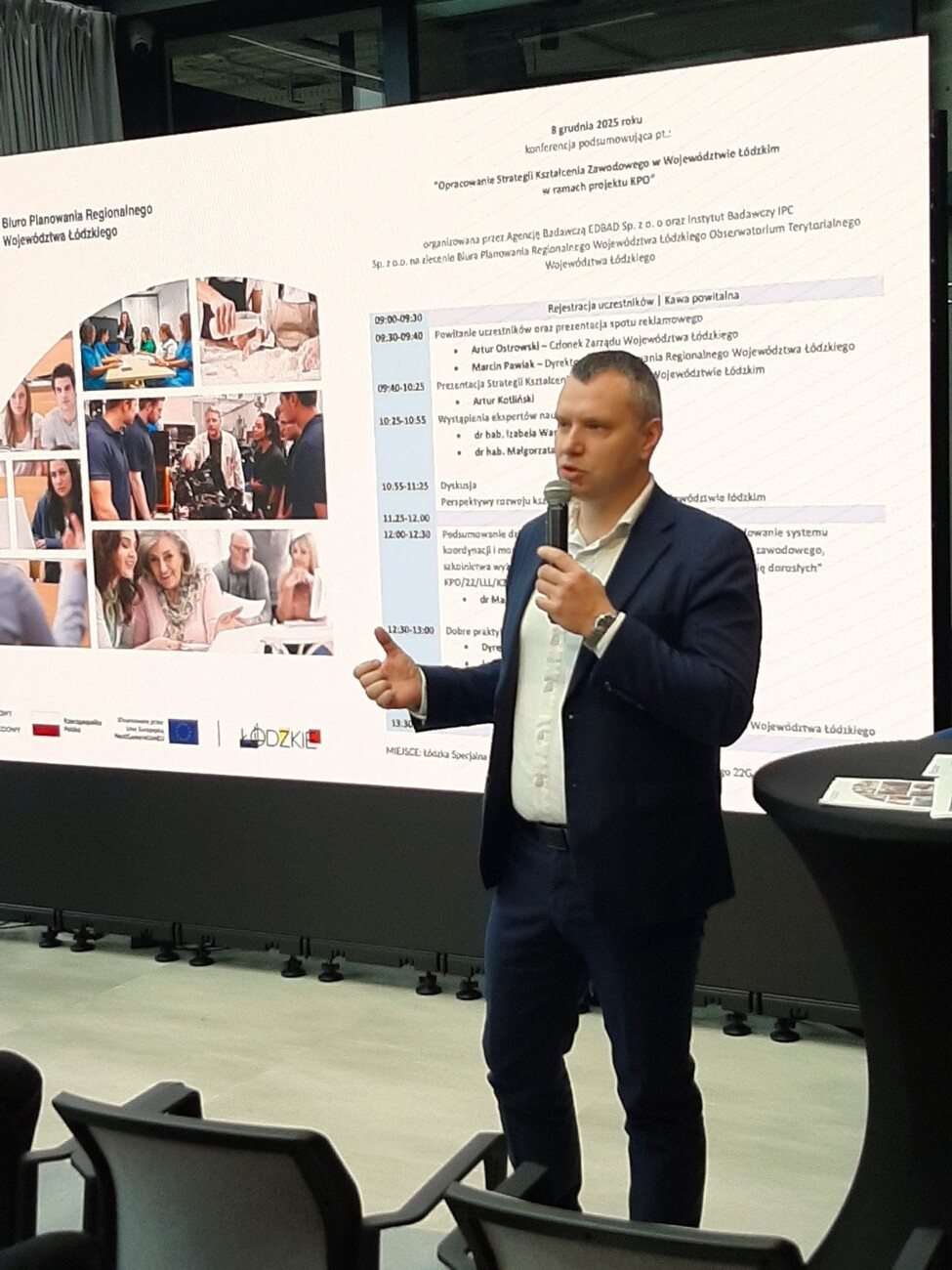 Na zdjęciu widać dyrektora Biura Planowania Regionalnego Województwa Łódzkiego Pana Marcina Pawlaka na tle telewizora wyświetlającego agendę konferencji.