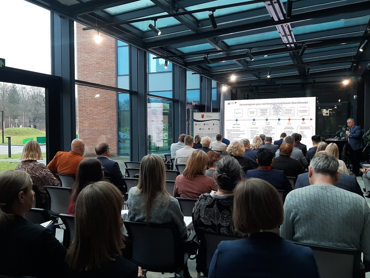 Zdjęcie przedstawia salę wraz z uczestnikami, na drugim planie widoczne jest na ekranie prezentacja dotyczącą Strategii Kstzałcenia Zawodowego Województwa Łodzkiego.